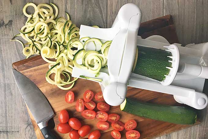 Horizontal image of a spiralizer with spiralized zucchini, chopped tomatoes, and a knife on a wooden cutting board. 