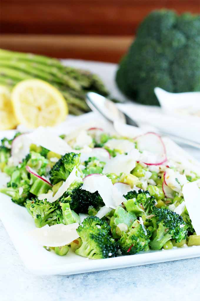 A serving dish of vegetable salad is in the foreground, with fresh lemon, broccoli, and asparagus in the background, with a white cloth and a serving spoon.