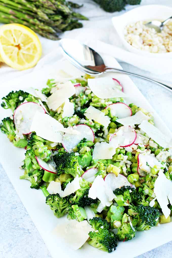Broccoli and asparagus salad, with radishes and parmesan cheese, arranged on a white serving platter beside a halved lemon, fresh vegetables, and a serving spoon on a speckled gray and white surface.
