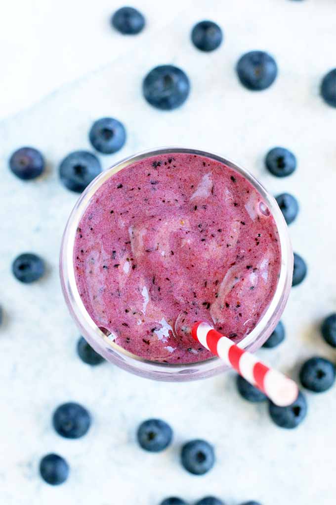 Top-down shot of a glass filled with a pinkish-purple fruit smoothie speckled with darker spots, with a white and red striped plastic straw, and fresh blueberries scattered on a light blue surface.