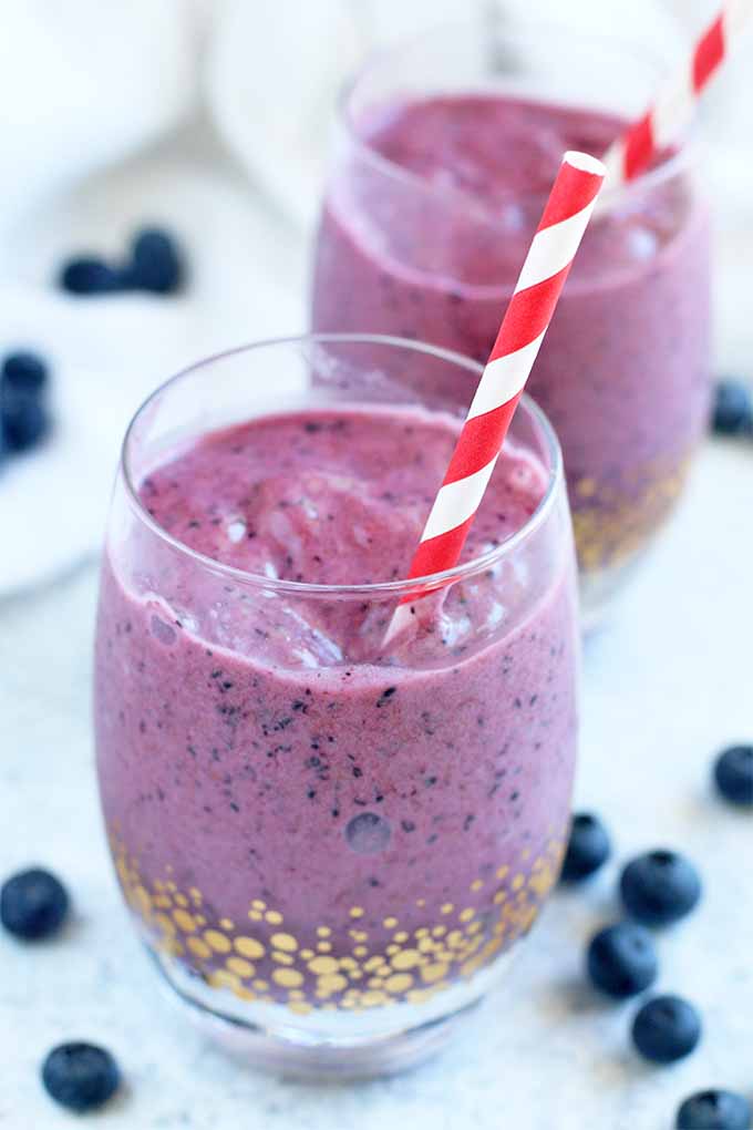 A glass filled with a purple fruit smoothie with a red and white striped straw, with another identical glass in the background, on a light blue surface with scattered whole blueberries.