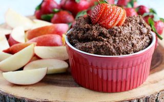 A red ramekin of dark chocolate hummus topped with a sliced strawberry for garnish, on a wood board topped with apple slices and more fresh berries in the background.