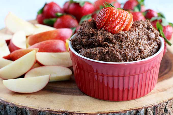 A red ramekin of dark chocolate hummus topped with a sliced strawberry for garnish, on a wood board topped with apple slices and more fresh berries in the background.