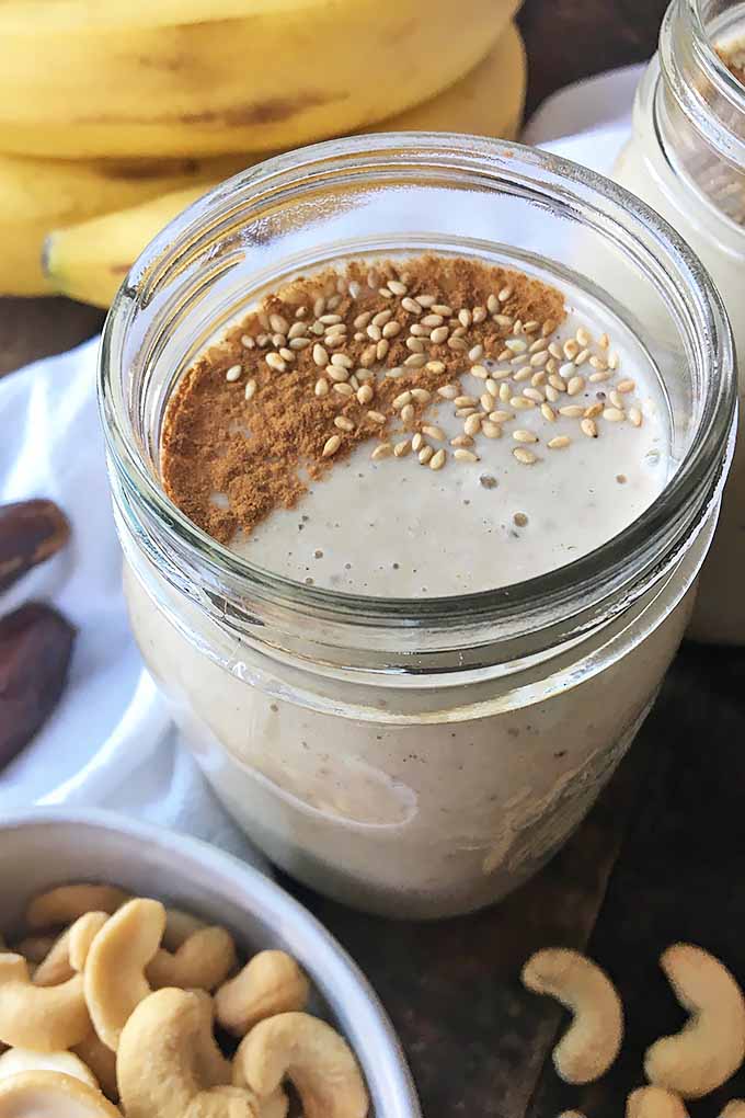 Vertical image of a jar glass with a light brown smoothie garnished with toasted sesame seeds and ground cinnamon. 