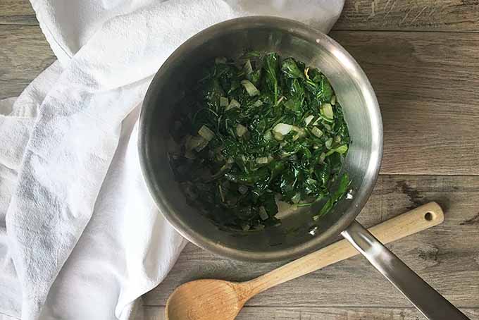 Horizontal image of a pot with sauteed greens and onions with a wooden spoon and a white towel.
