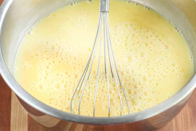 A large stainless steel mixing bowl of pale orange sherbet base being stirred with a whisk, on a brown wood surface.