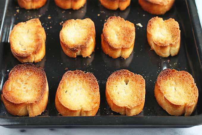 Toasted baguette rounds arranged in rows on a black rimmed baking sheet.