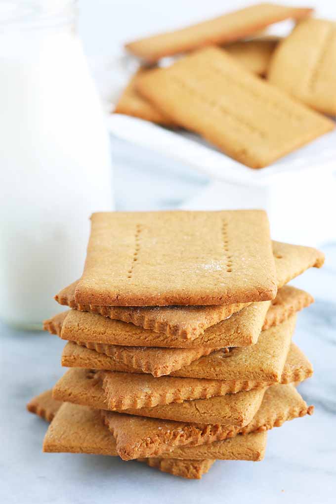 A tall stack of homemade graham crackers is in the foreground, with a small glass bottle of milk, and more of the baked goods in soft focus in the background, on a gray and white marble surface.