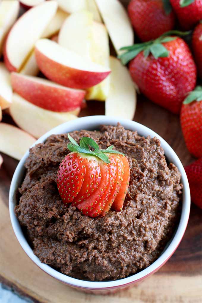 A small dish is filled with chocolate hummus and topped with a sliced strawberry, with more strawberries and red apple slices on a wooden surface.