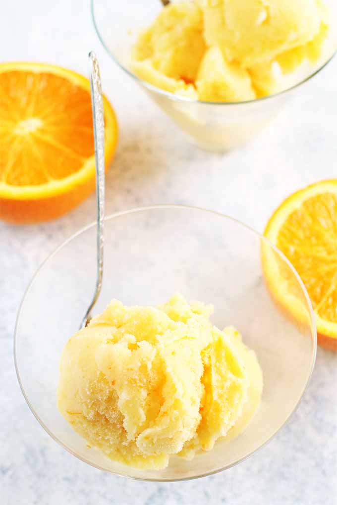 Top-down vertical image of two clear parfait glasses of scoops of sherbet with a silver spoon, on a white sponge painted surface with two halves of an orange, arranged cut side up.