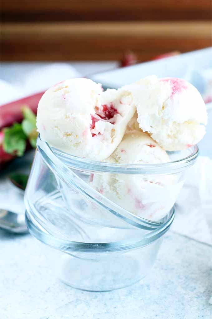 Three scoops of rhubarb swirl ice cream in the topmost clear glass dish in a stack of three, with spoons and stalks of rhubarb in the background, on a blue and white speckled countertop with a wooden wall in shallow focus.