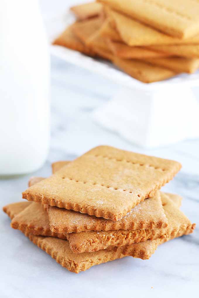 Vertical image of a stack of golden brown freshly baked graham crackers, next to a bottle of milk and a white square cake stand displaying more of the baked good, on a marble surface.