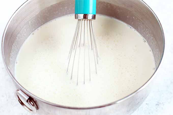 Closeup of a blue-handled whisk stirring sugar, milk, and cream in a stainless steel mixing bowl.