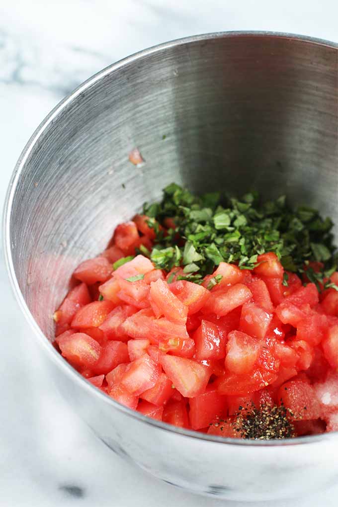 A stainless steel mixing bowl that contains chopped tomatoes and basil, salt, and pepper, on a white marble surface with gray veins.