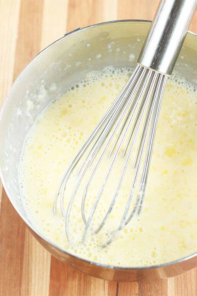 Closeup of a wire whisk stirring a yellow foamy mixture in a large stainless steel mixing bowl, on a striped wood background.