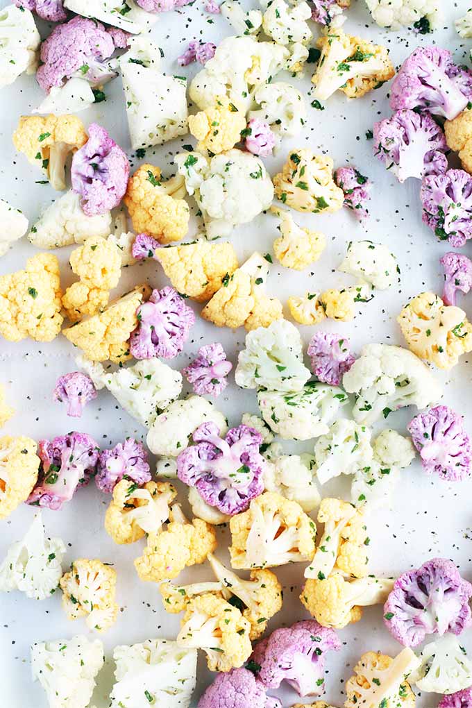 Top-down image of orange, purple, and white raw cauliflower florets, coated with olive oil and minced fresh herbs, on a white parchment background.