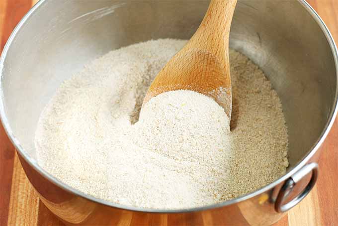 Closeup of a wooden spoon stirring spelt flour and other dry ingredients in a large stainless steel mixing bowl with a ring handle, on a wood background.