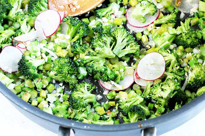 A frying pan full of chopped green vegetables, thinly sliced radishes, and lemon zest, being stirred with a wooden spoon.