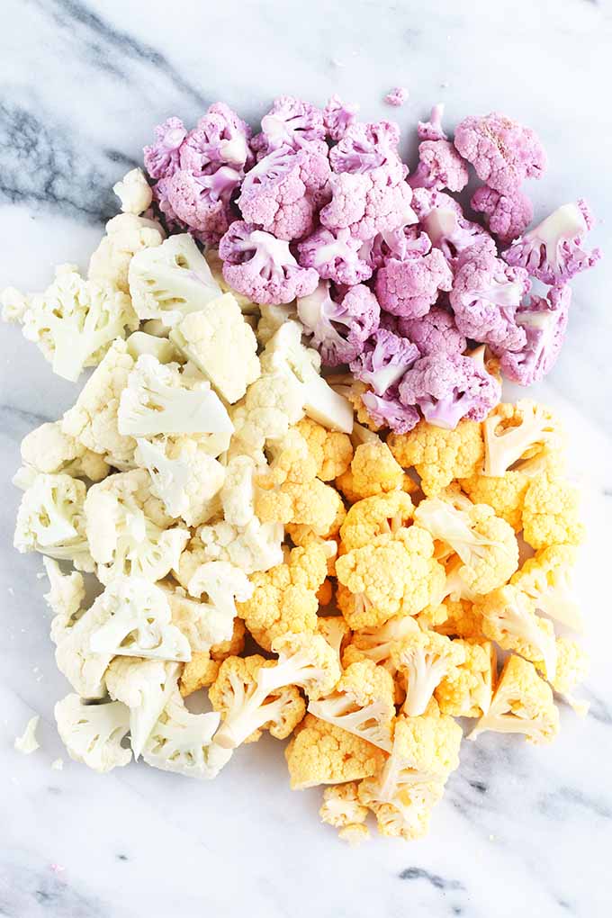 Chopped purple, orange, and white cauliflower florets arranged in a pile on a white piece of marble with gray veins.