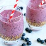 Two glasses with a gold decorative pattern at the base, filled with a purple-colored fruit smoothie, on a light blue surface covered with scattered fresh blueberries, with red and white straws stuck into the drinks.