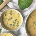 Horizontal image of three bowls of creamy potage on a white towel with fresh spinach leaves and a wooden spoon.