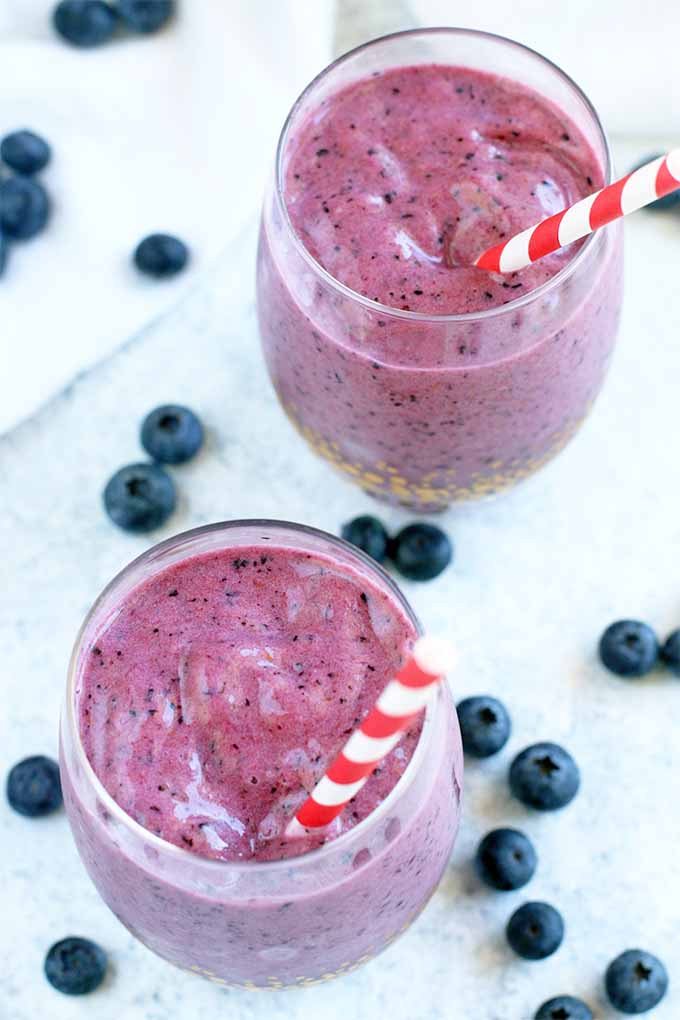 Top-down view of two glasses filled with a pinkish-purple fruit smoothie, with scattered blueberries.