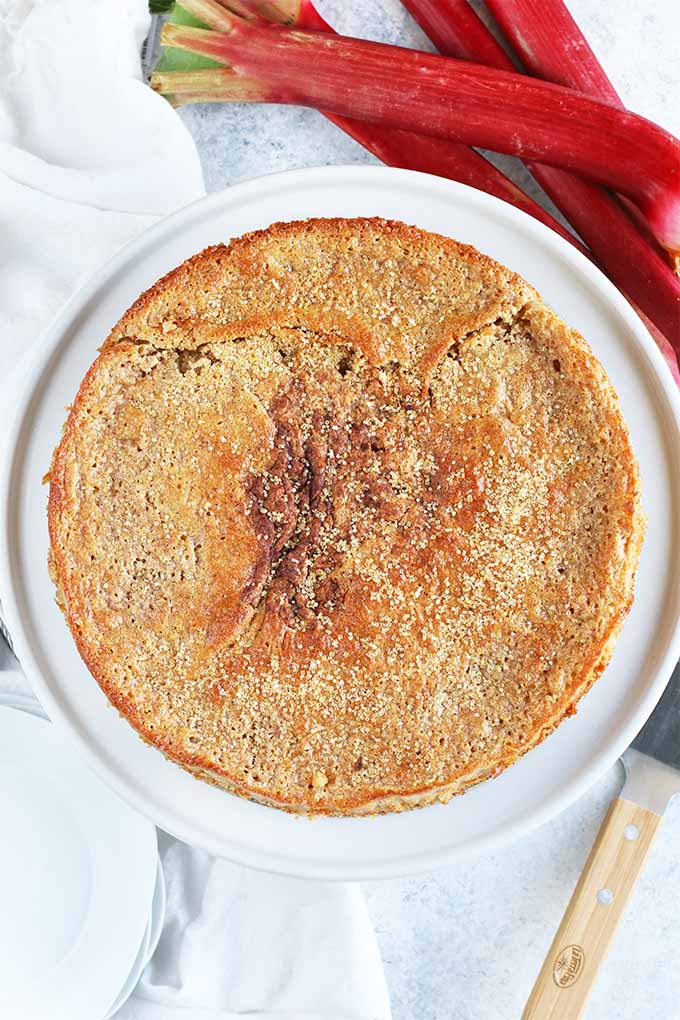 Top-down image of a round, golden brown spelt cake topped with turbinado sugar, on a white cake stand with a white cloth and several stalks of red rhubarb alongside a wood and metal cake server.