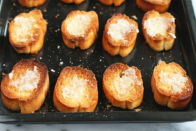 Toasted baguette slices arranged in rows on a black rimmed baking sheet, with garlic paste spread on top.
