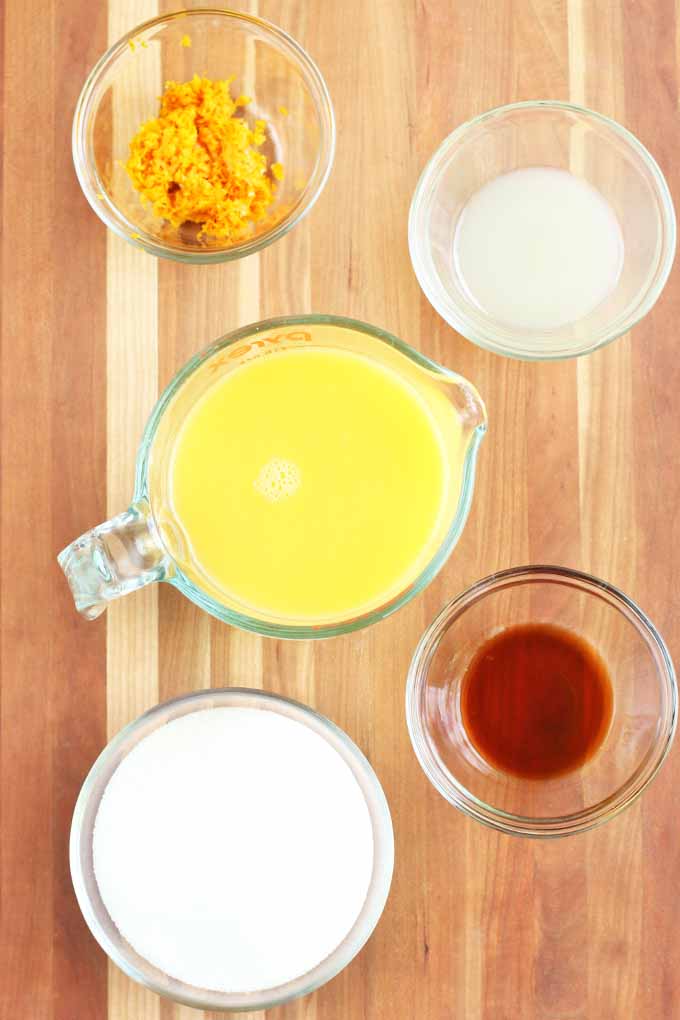 Top-down vertical image of small glass dishes or orange zest, sugar, vanilla extract milk, and a glass pitcher of orange juice, on a wooden surface with vertical stripes.