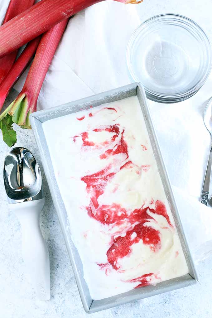 Top-down view of a metal loaf pan filled with white ice cream with a red swirl, with a white plastic and metal ice cream scoop, glass dessert dishes, a white cloth napkin, and stalks of pink rhubarb with green tips.
