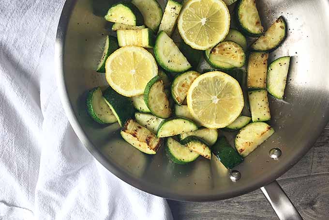 Horizontal image of a skillet with cooked zucchini and lemons on a white towel.