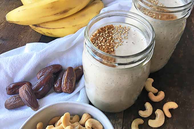 Horizontal image of jar glasses filled with a light smoothie mixture surrounded by cashews, dates, bananas, and a white napkin on a wooden surface.