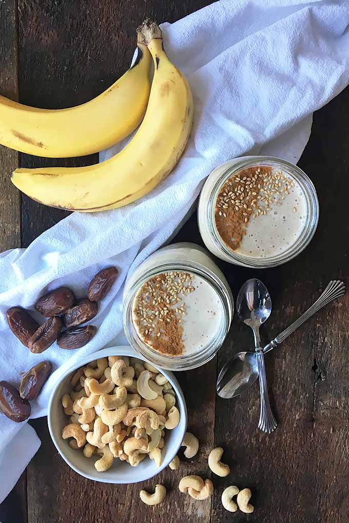 Vertical image of two garnished smoothies, spoons, cashews, dates, and bananas