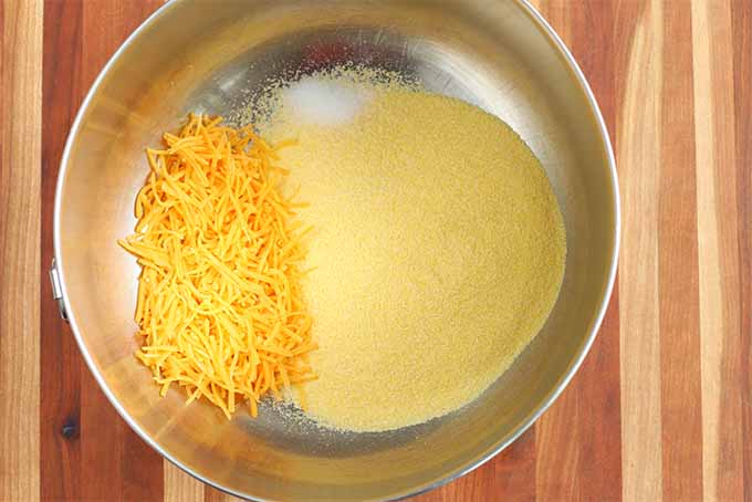 Top-down shot of a stainless steel bowl of yellow cornmeal, salt, and orange shredded cheddar cheese, on a brown wood background.