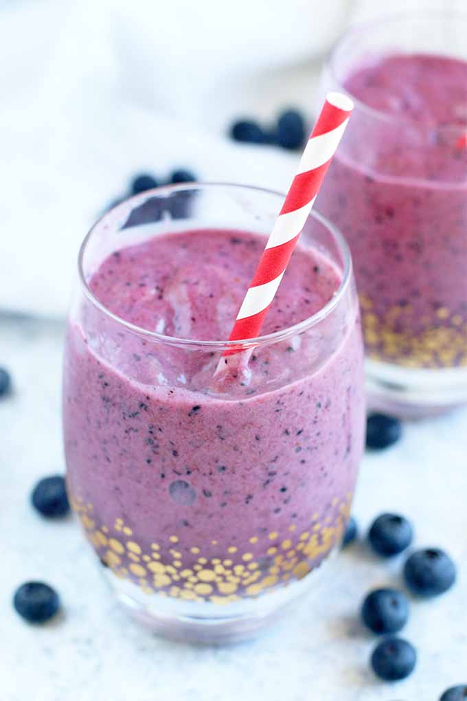Two glasses with gold decoration, filled with blueberry orange banana smoothie, with red and white striped straws and blueberries scattered on a light blue surface.