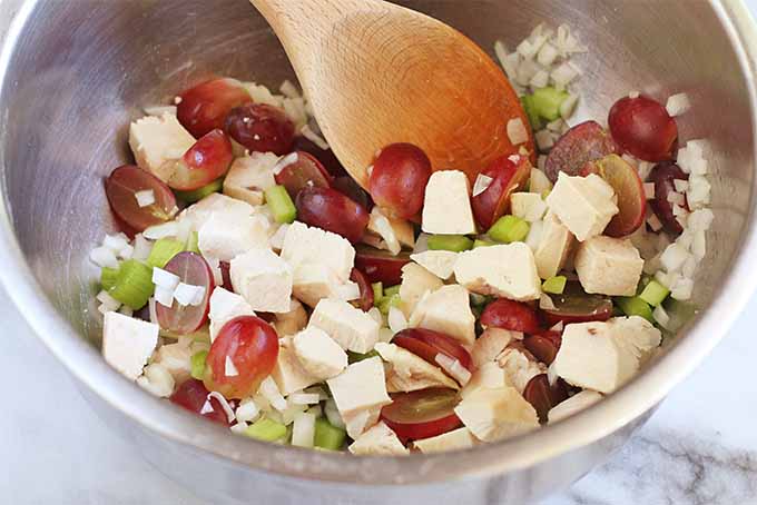 Top-down view of a mixture of red grapes, celery, cooked chicken, and onion, in a stainless steel m mixing bowl being stirred together with a wooden spoon.
