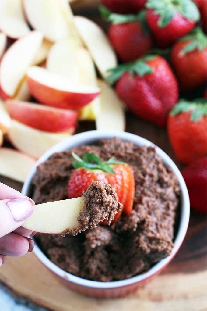 A hand with pink manicured nails holds a slice of apple dipped into a red and white ramekin of chocolate hummus topped with a sliced strawberry, on a wooden board with more fresh apple slices and whole strawberries.