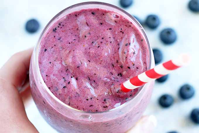 Top-down view of a hand holding a glass of pinkish-purple blueberry smoothie, with a red and white striped straw, and scattered blueberries on a light blue background.
