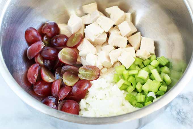 Top-down view of a stainless steel mixing bowl filled with four separate piles of bite-sized cooked white chicken breast, chopped celery, chopped onion, and halved red grapes.
