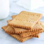A small stack of five homemade graham crackers with scored edges and rows of holes in the top of each, beside a glass bottle of milk and a white ceramic cake stand, on a gray and white marble background.