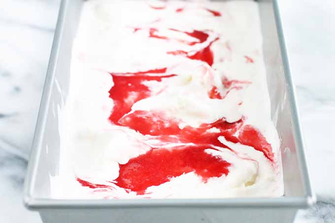A stainless steel loaf pan filled with homemade vanilla ice cream swirled with rhubarb sauce, on a gray and white marble background.