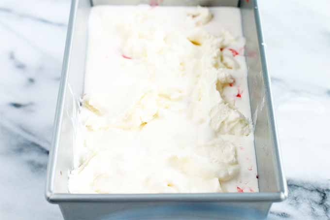 A metal baking pan filled with vanilla ice cream swirled with rhubarb sauce, on a white and gray marble background.