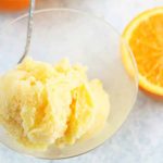 A scoop of orange sherbet in a glass bowl with a spoon, on a white and pale blue sponge painted surface with two halves of a cut orange.