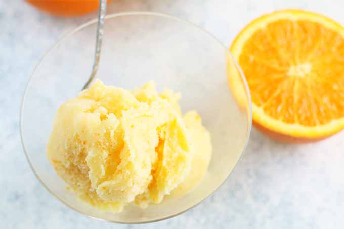 A scoop of homemade sherbet in a glass bowl with a spoon, on a white and pale blue sponge painted surface with two halves of a cut orange.