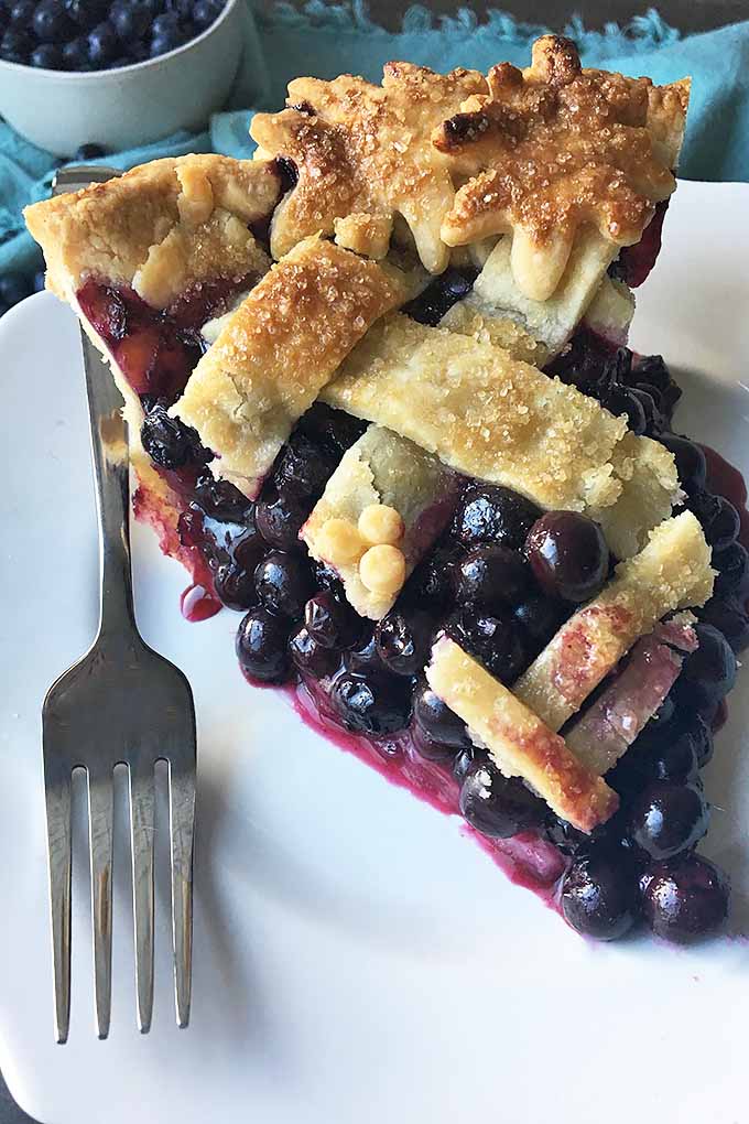 Vertical image of a slice of blueberry pie with a fork on a white plate. 