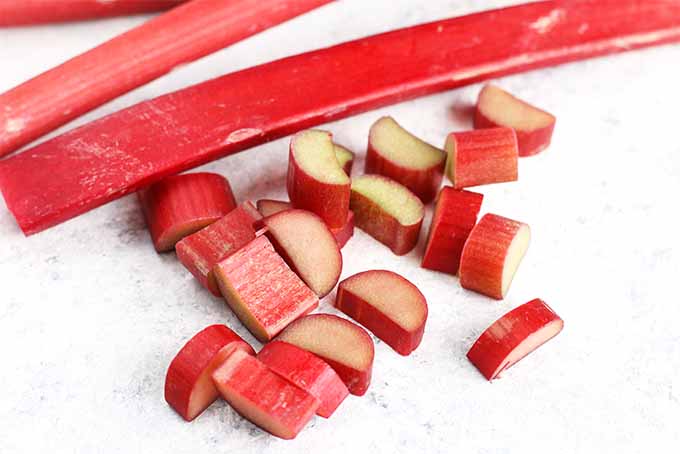Closeup of a few whole stalks of pinkish-red rhubarb with a small pile of chopped pieces that are pale green on the inside and red on the outside, on a blue speckled white background.