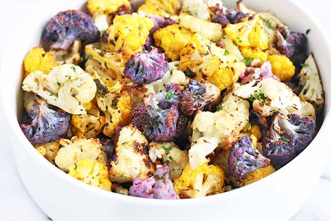 A white serving dish of chopped, roasted, multicolored cauliflower with fresh herbs, on a white marble background.