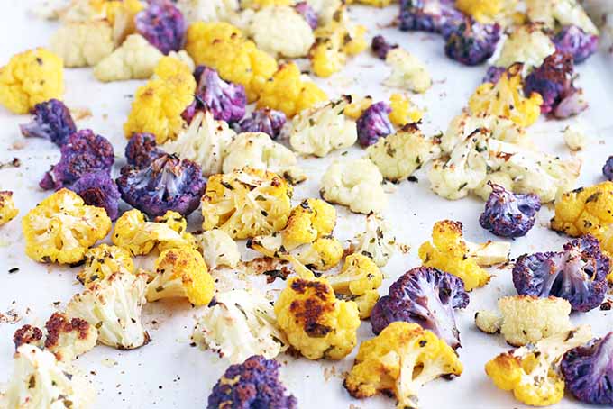 Closeup of orange, white, and purple roasted cauliflower florets coated with chopped fresh herbs, on a white parchment paper background.