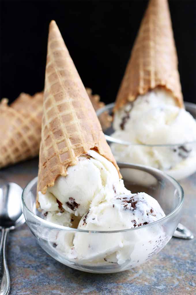 Two glass bowls of vanilla and chocolate stracciatella ice milk, with inverted waffle cones on top and more resting on the granite countertop behind them with a few scattered spoons, on a black background.
