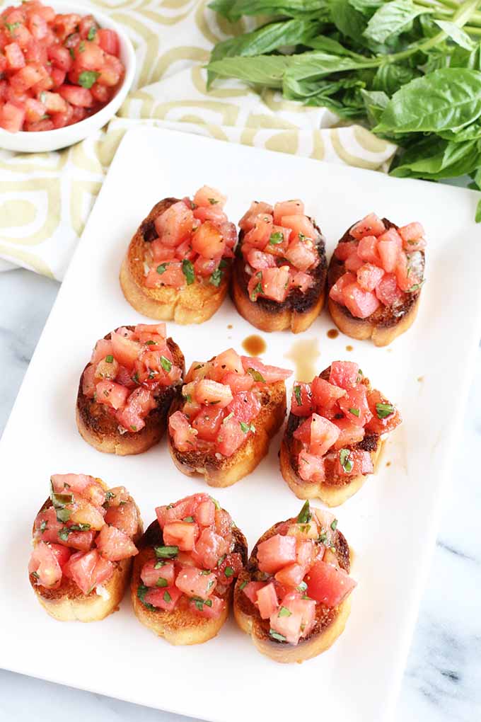 Oblique view of nine pieces of bruschetta on a square serving platter, on a gray marble surface with a folded cloth napkin, a small white bowl of chopped tomatoes, and a bunch of basil.